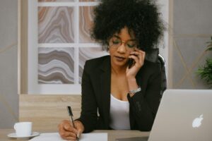 woman in black blazer on the phone while taking notes at a desk with an open laptop